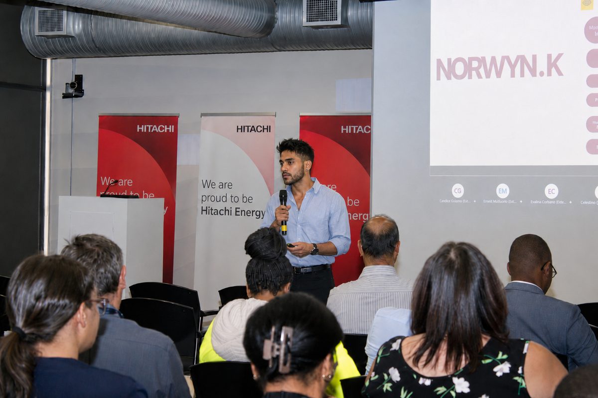 Norwyn K speaking with a microphone to a seated audience at a corporate event.